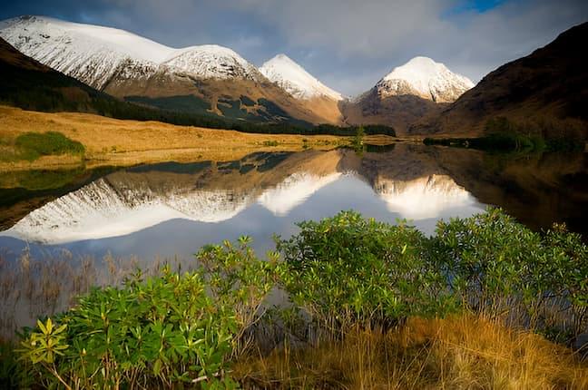 view of snow-capped mountains in Scotland, reflected in a calm lake, with lush green foliage and grassy terrain in the foreground.
