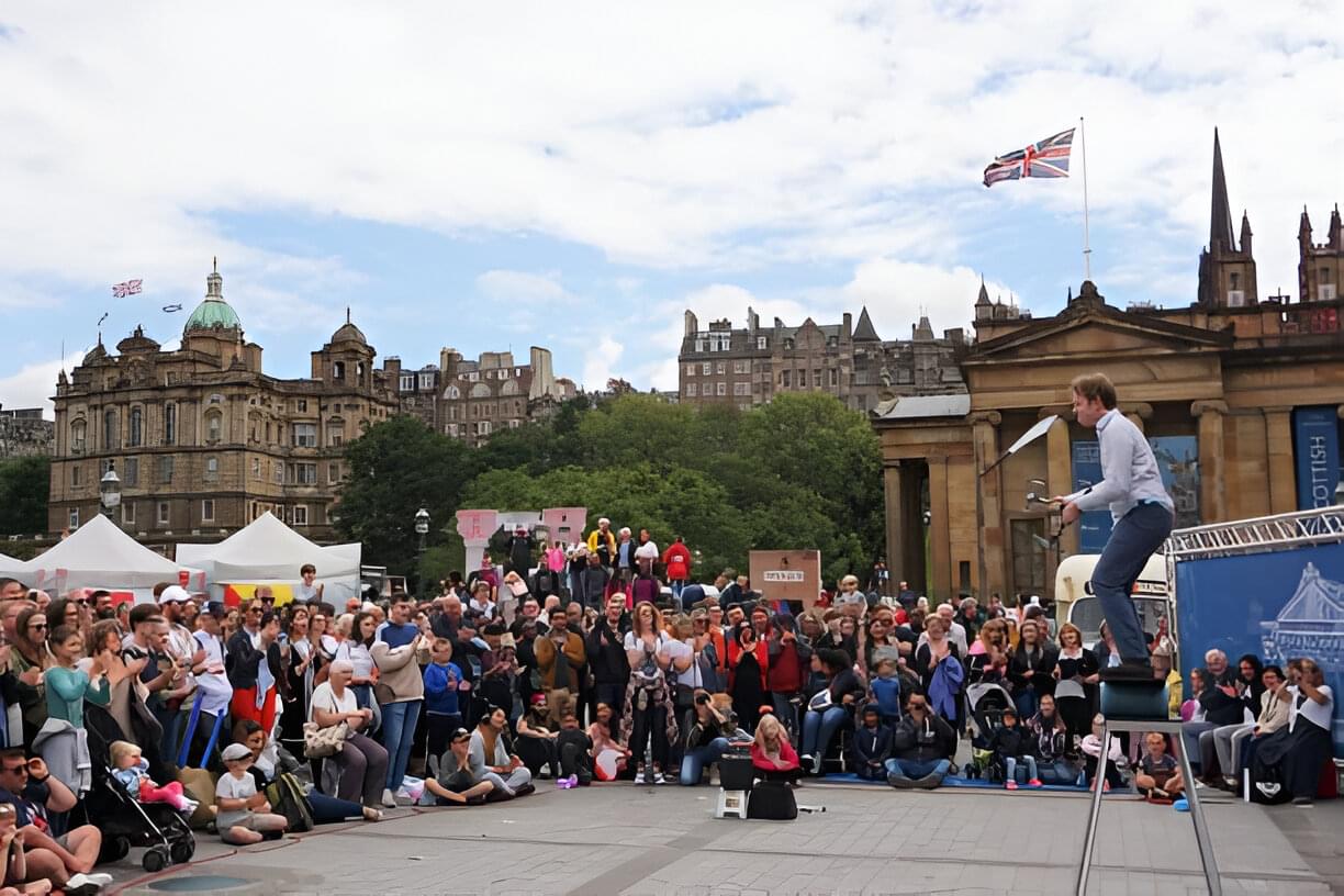 A street performance in Edinburgh with a juggler performing on a stool. The backdrop features historic buildings.