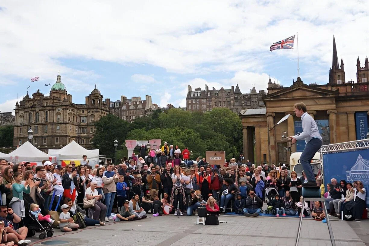 A street performance in Edinburgh with a juggler performing on a stool. The backdrop features historic buildings.