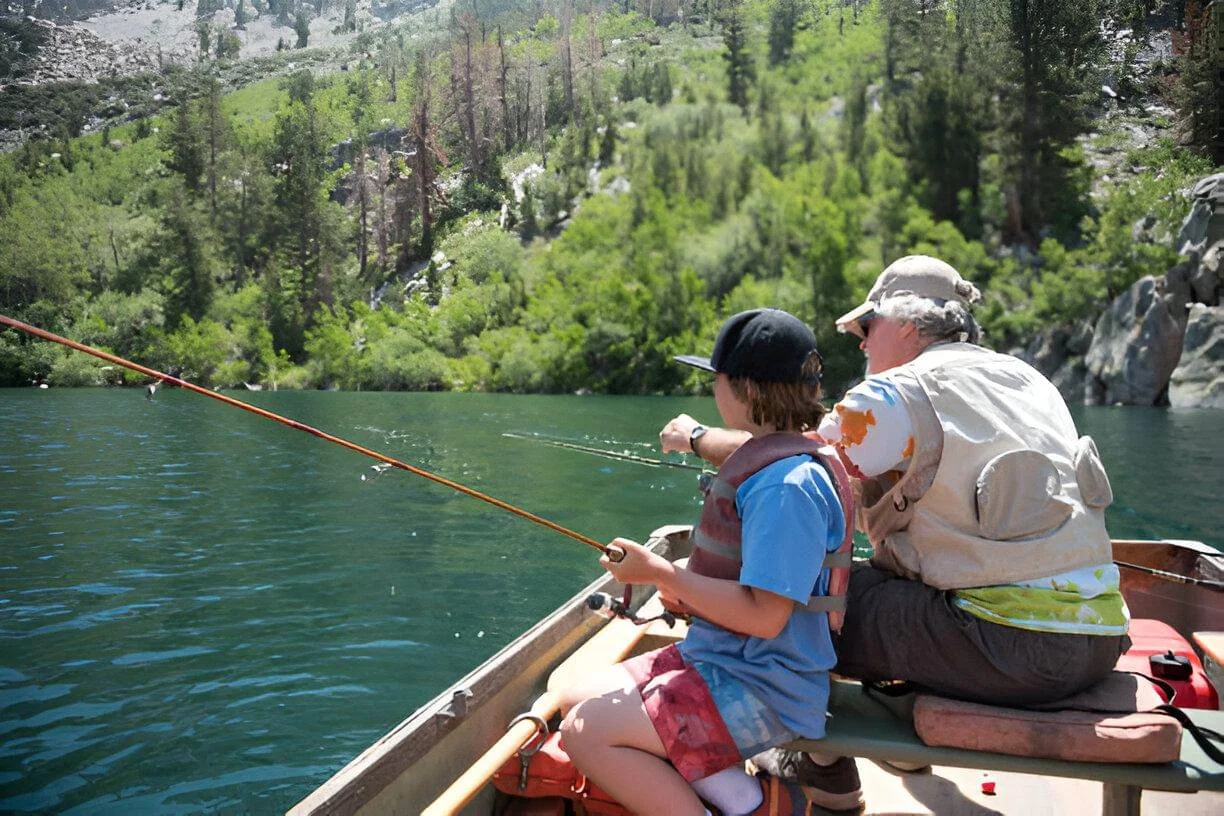 A child and adult fishing together on a boat in a lake, surrounded by greenery and mountains.
