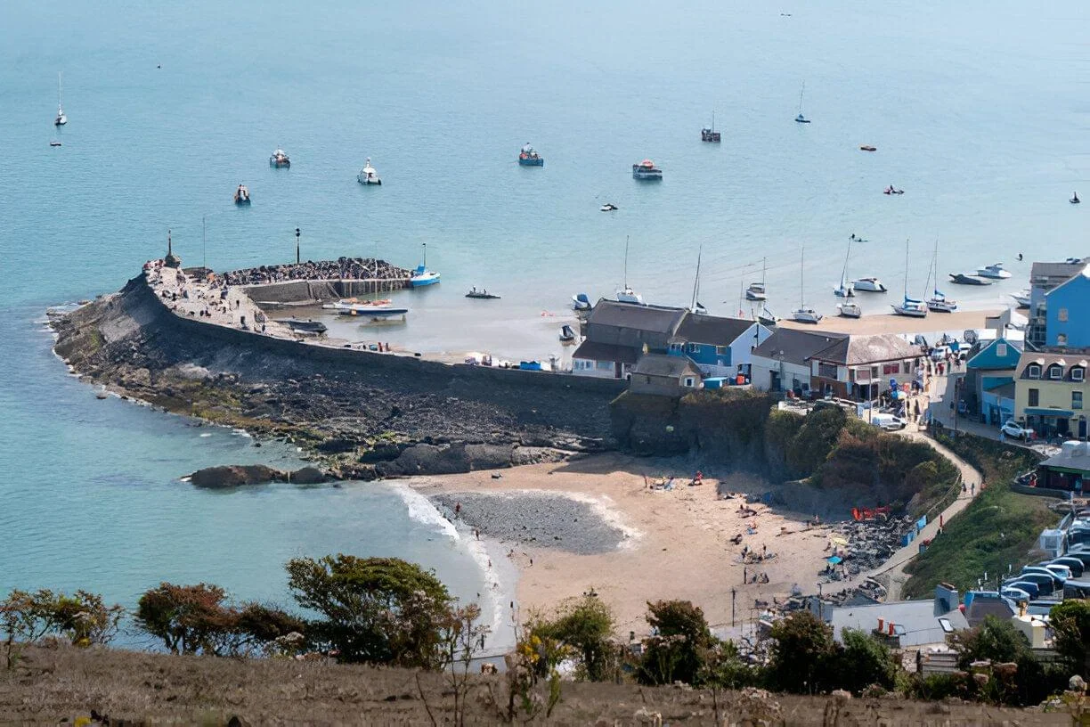 A scenic view of New Quay, Wales, showing a small harbor with boats, a sandy beach with people enjoying the sun.
